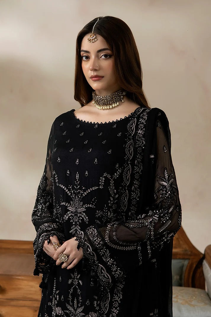 A woman wearing a black traditional embroidered outfit with sheer sleeves and ornate silver-toned detailing, styled with a statement necklace and headpiece against a softly lit beige interior background.
