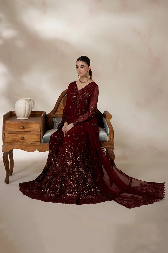 A woman seated on a wooden chair wearing a deep maroon embroidered traditional gown with a matching sheer draped overlay, posed in a studio with a beige backdrop and a small side table holding a white vase.