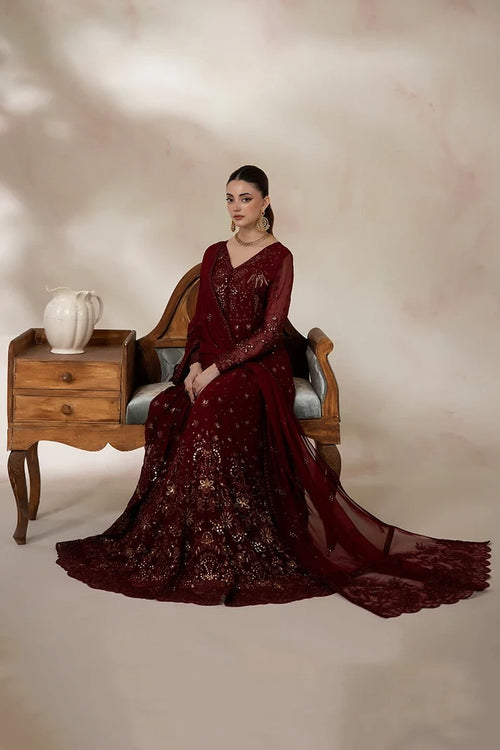 A woman seated on a wooden chair wearing a deep maroon embroidered traditional gown with a matching sheer draped overlay, posed in a studio with a beige backdrop and a small side table holding a white vase.