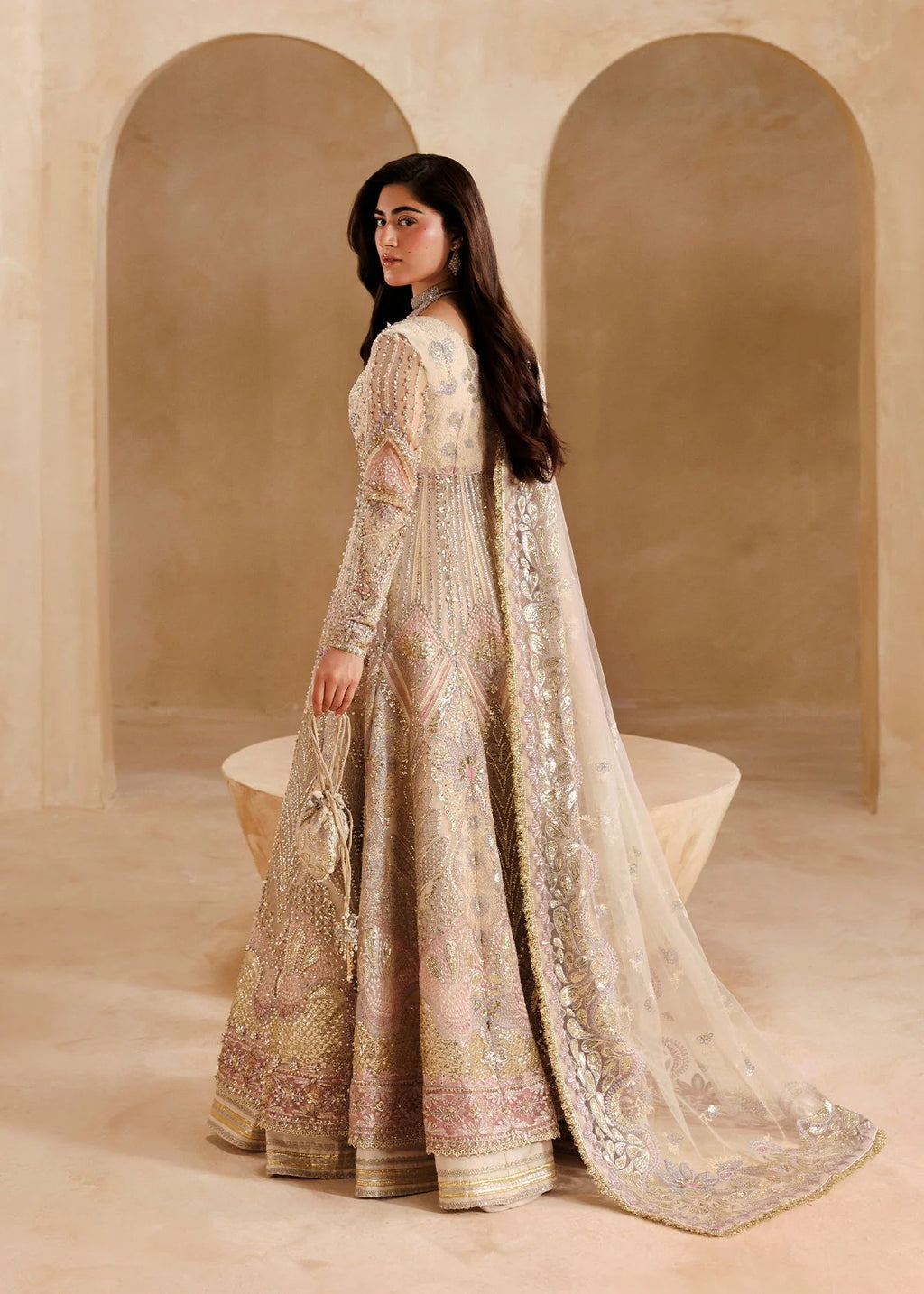 A woman wearing an ornate beige and gold embroidered traditional gown with a long sheer dupatta, standing in a warm-toned architectural setting with arched alcoves.
