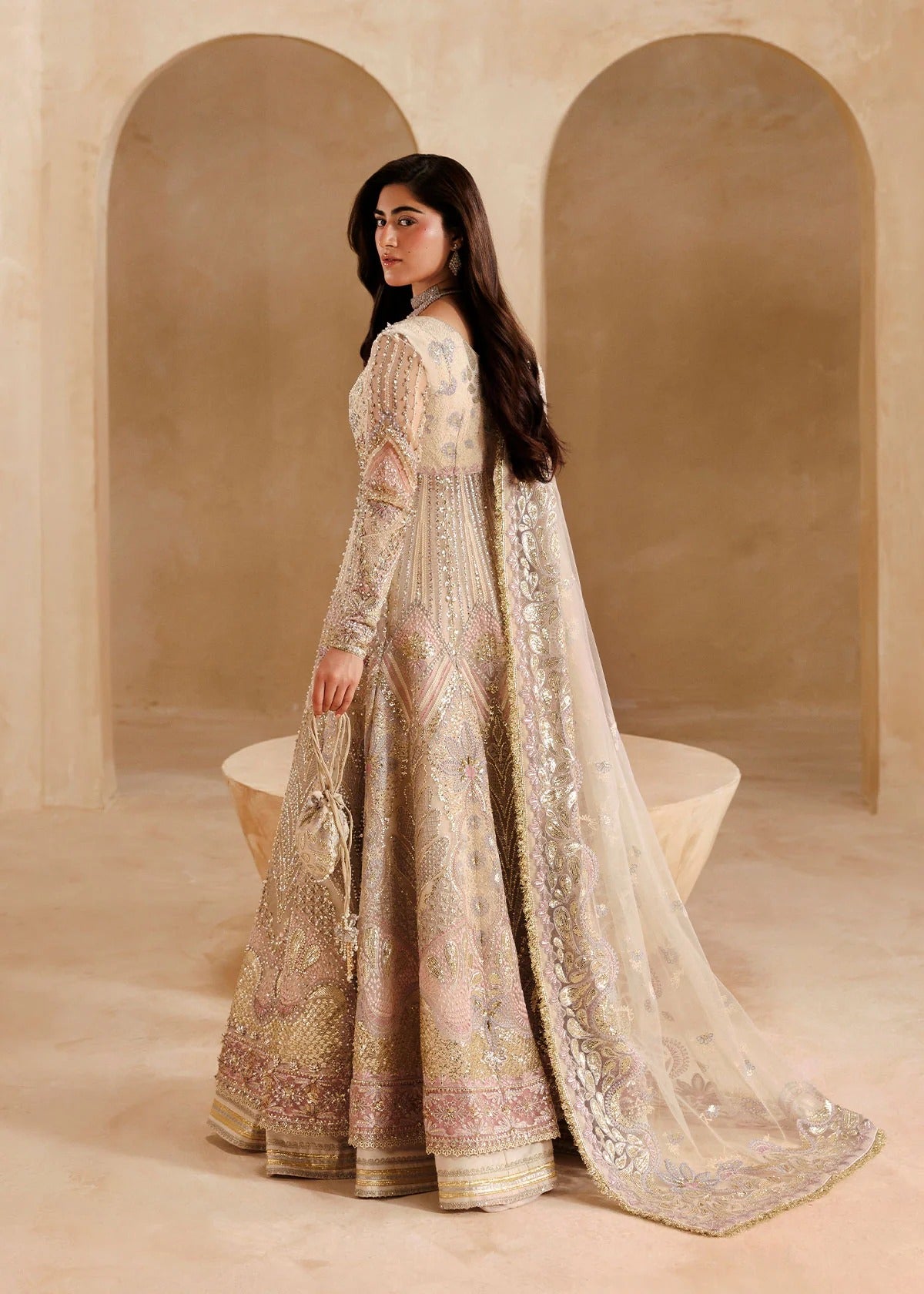 A woman wearing an ornate beige and gold embroidered traditional gown with a long sheer dupatta, standing in a warm-toned architectural setting with arched alcoves.