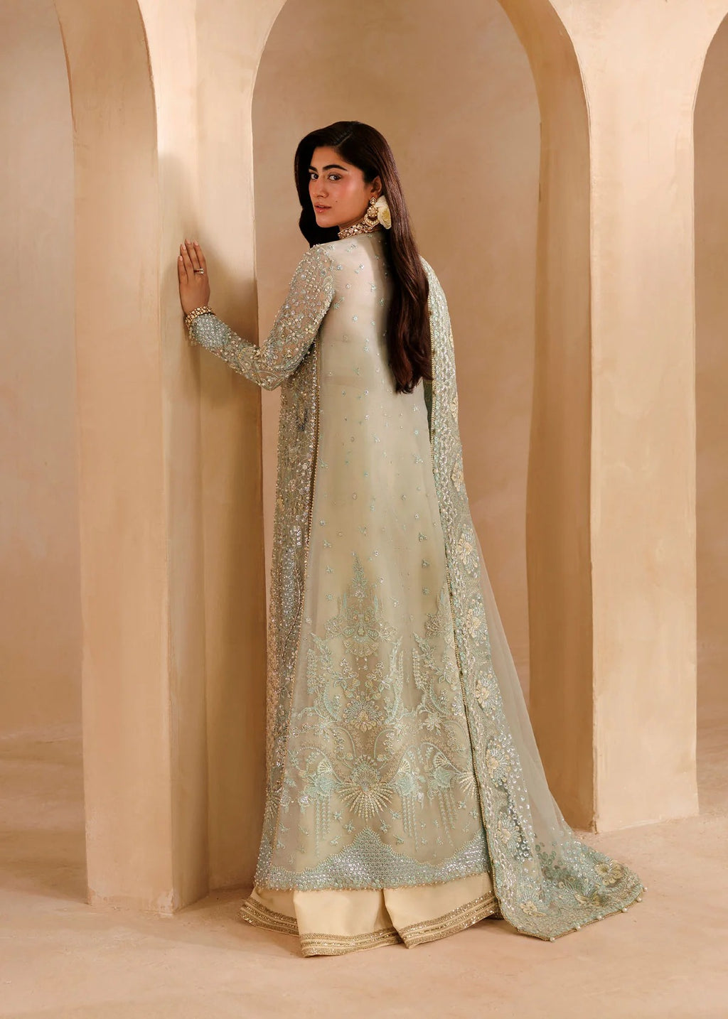 A woman wearing a traditional Indian outfit with a light sage green embroidered kurta, matching dupatta, and beige trousers, posing in front of beige arches.