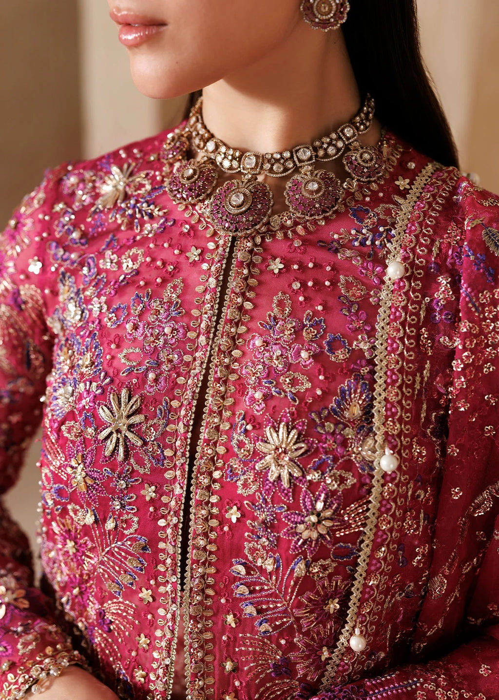 A close-up of a model wearing a heavily embellished magenta pink traditional embroidered jacket with intricate gold, silver, and purple beadwork and embroidery.