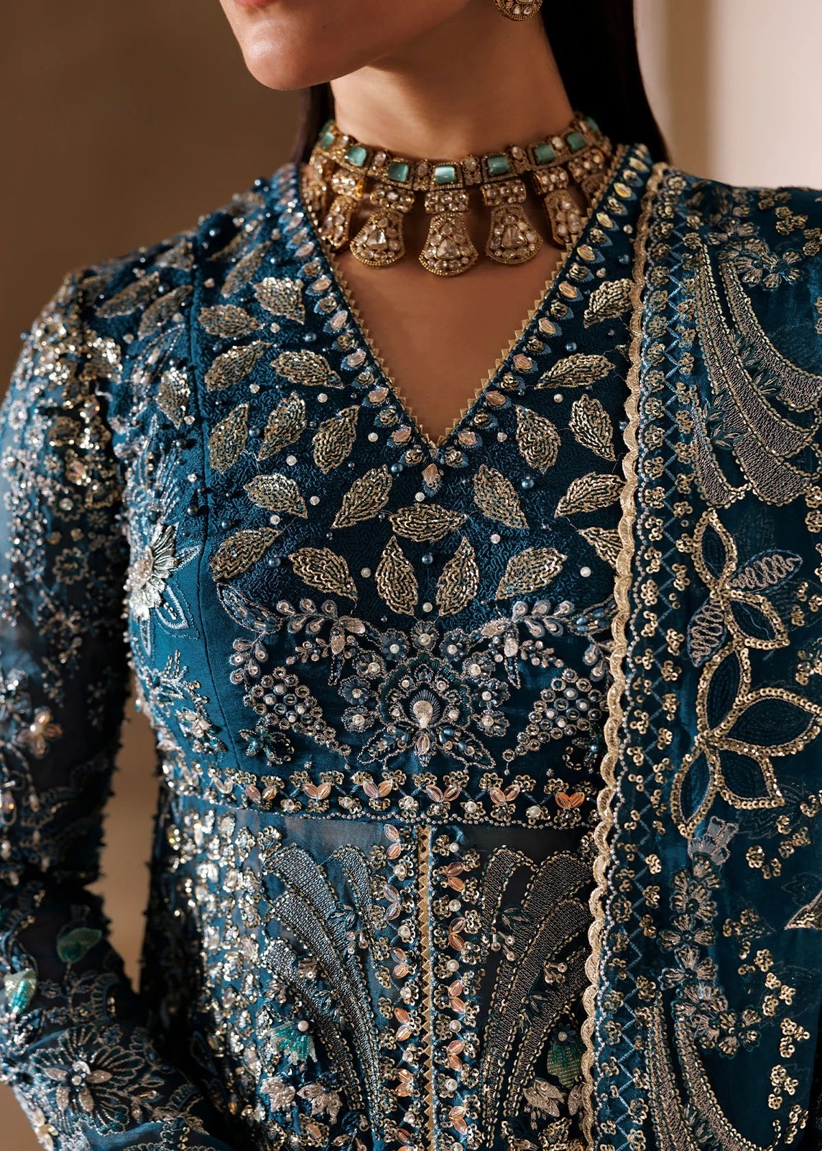 Close-up of a model wearing an intricately embroidered teal blue traditional outfit with gold and silver beadwork, a matching embroidered dupatta, and a statement gold choker necklace with turquoise accents.