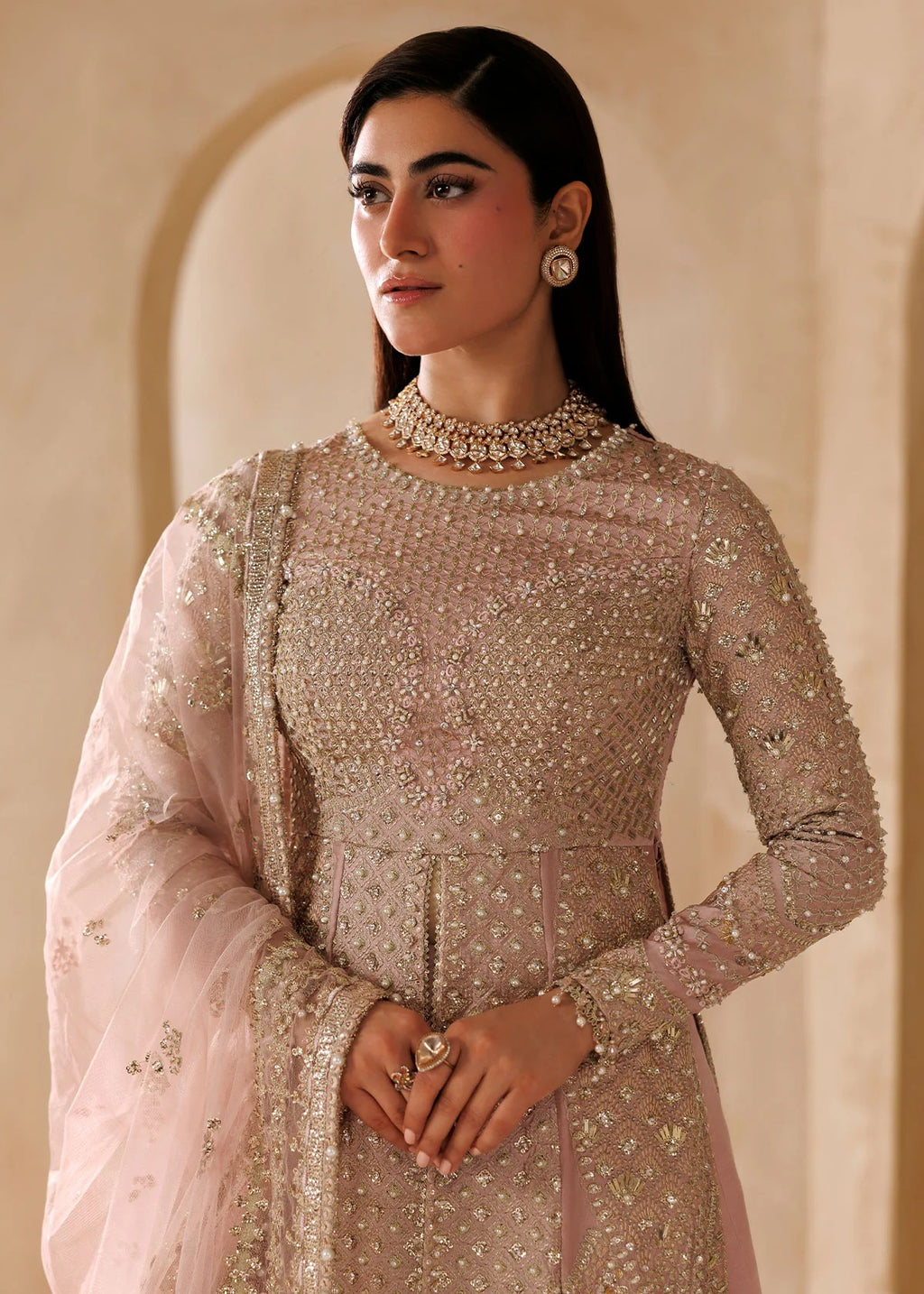 A woman wearing an embellished light pink traditional outfit with a matching sheer dupatta, standing against a warm beige architectural background.