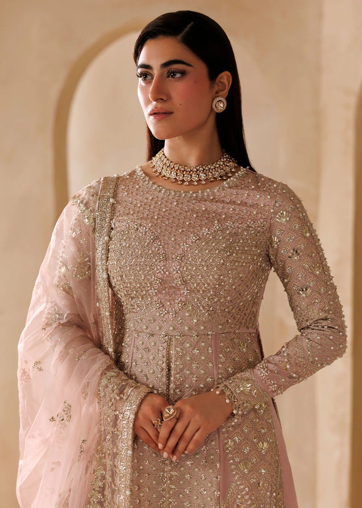 A woman wearing an embellished light pink traditional outfit with a matching sheer dupatta, standing against a warm beige architectural background.