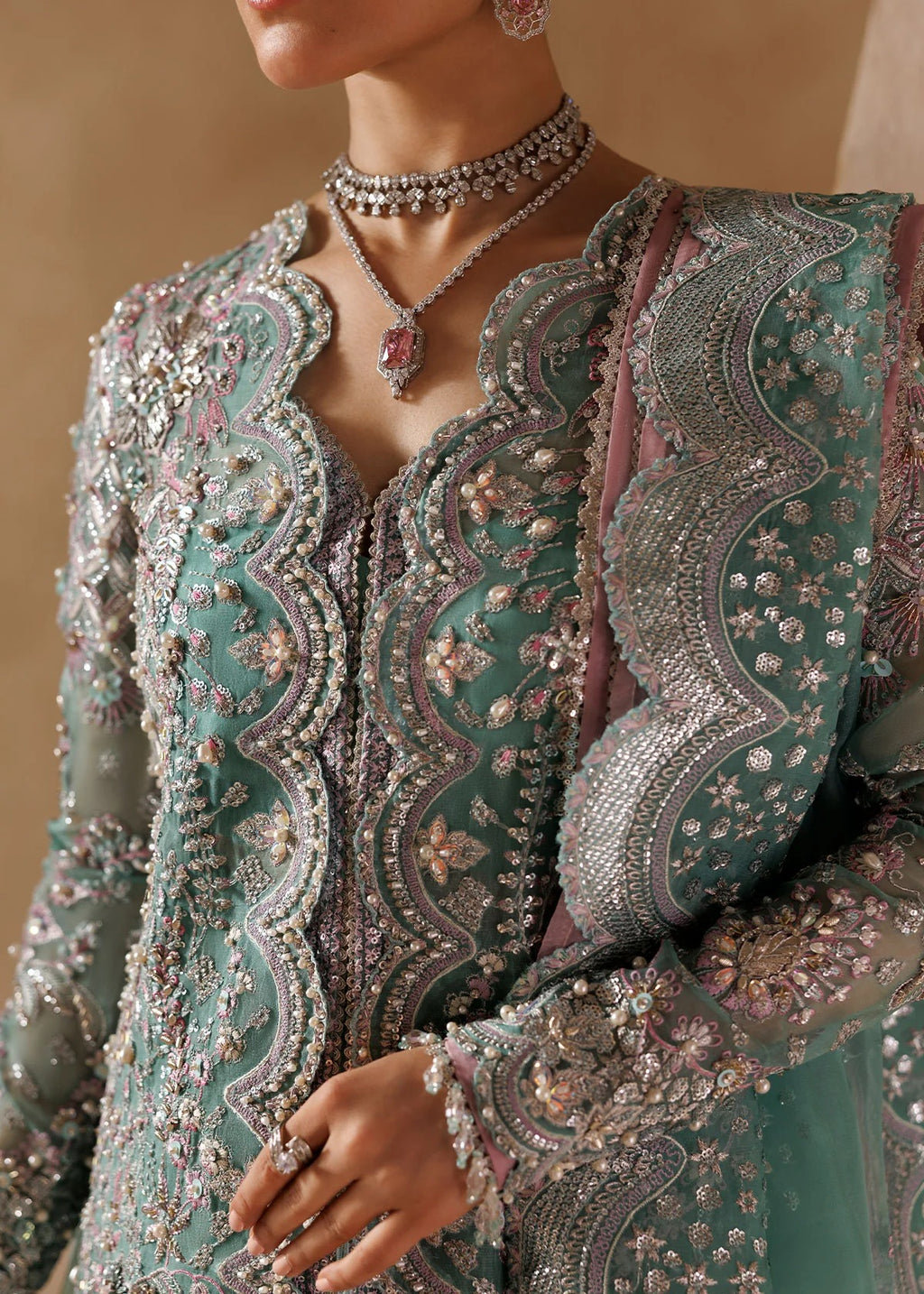 A close-up of a model wearing an intricately embroidered traditional outfit in dusty teal green with silver, pink, and white beadwork, paired with a matching sheer dupatta and jewelry.