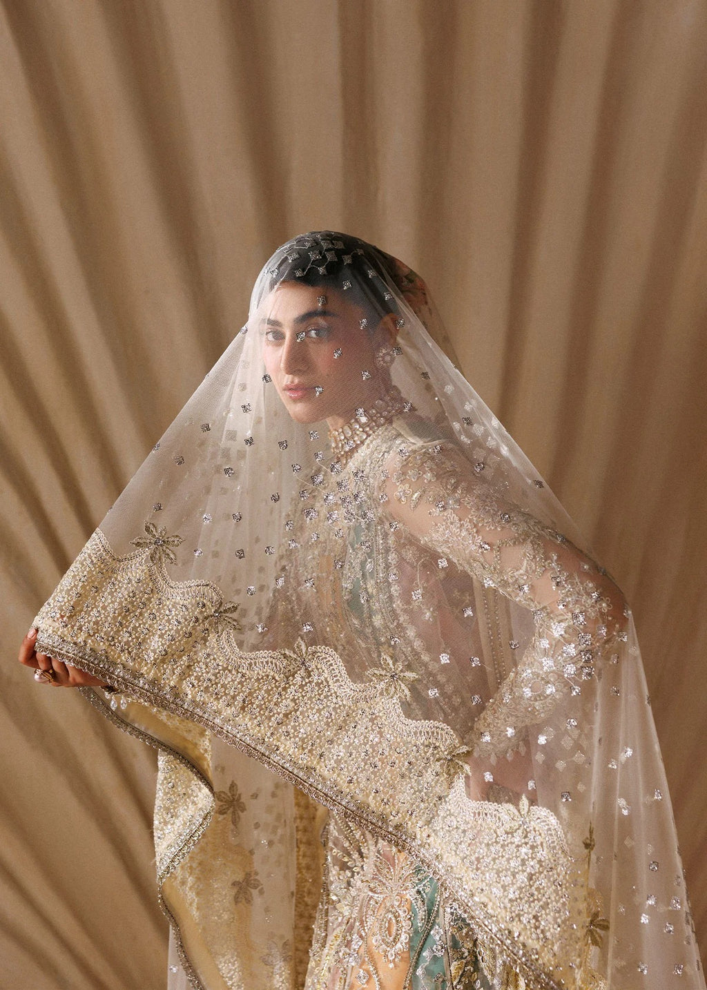 A woman wearing an ornate cream and gold embellished traditional bridal outfit with a sheer embroidered veil, posed against a draped beige fabric backdrop.