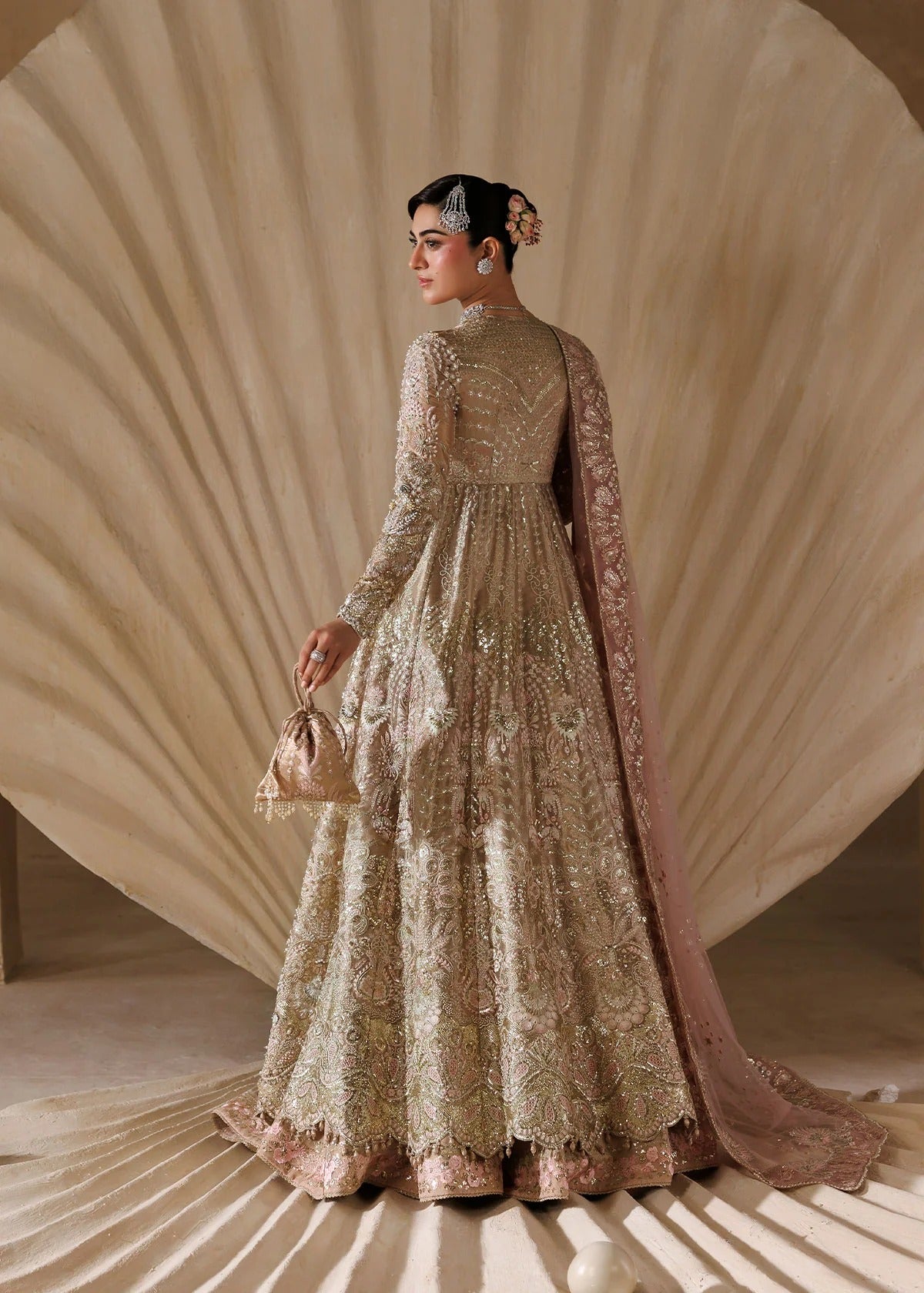 A woman stands in profile wearing an ornate beige and blush pink embroidered traditional gown with a long draped overlay, holding a small matching handbag against a sculptural beige backdrop.