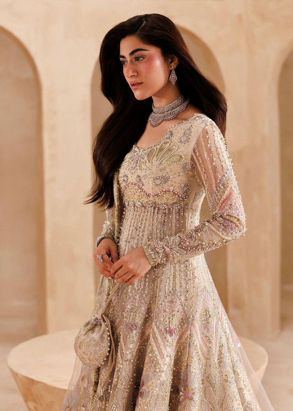 A woman wearing a heavily embellished beige and gold traditional gown with long sheer sleeves, standing in a warm-toned architectural setting with arches.