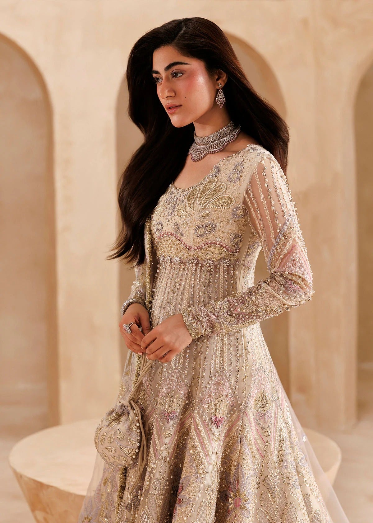 A woman wearing a heavily embellished beige and gold traditional gown with long sheer sleeves, standing in a warm-toned architectural setting with arches.