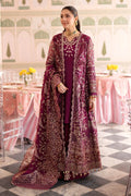 A woman models a traditional deep magenta embroidered outfit with a matching sheer jacket in an ornate indoor setting with pink tables and a checkered floor.