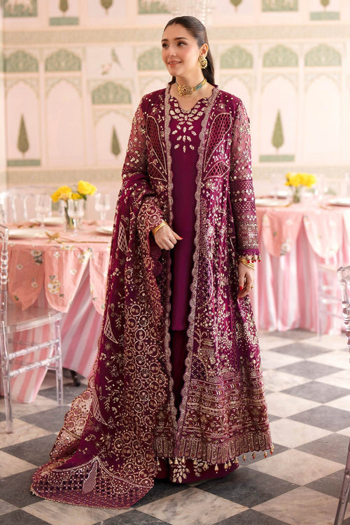 A woman models a traditional deep magenta embroidered outfit with a matching sheer jacket in an ornate indoor setting with pink tables and a checkered floor.