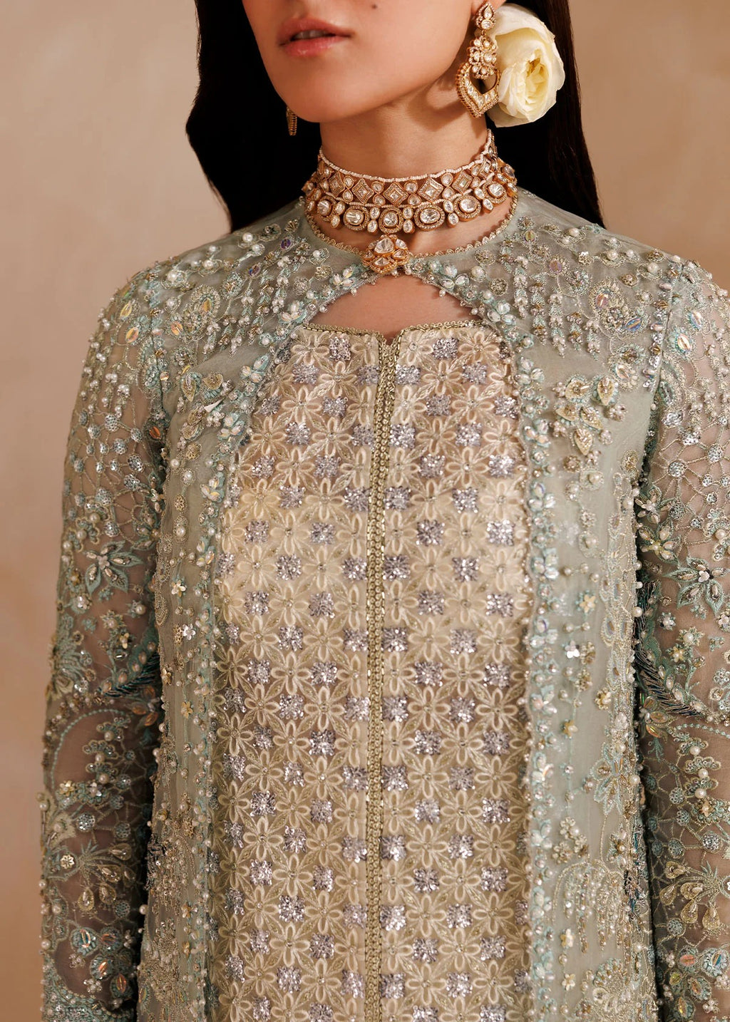 A close-up of a model wearing an ornate traditional outfit with intricate silver and gold embroidery on a light sage green and beige fabric, accessorized with a detailed gold choker necklace and a large cream flower earring.