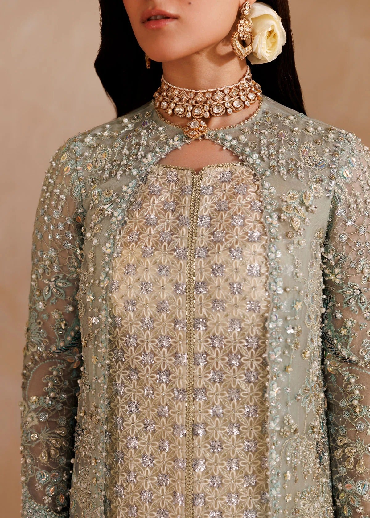 A close-up of a model wearing an ornate traditional outfit with intricate silver and gold embroidery on a light sage green and beige fabric, accessorized with a detailed gold choker necklace and a large cream flower earring.