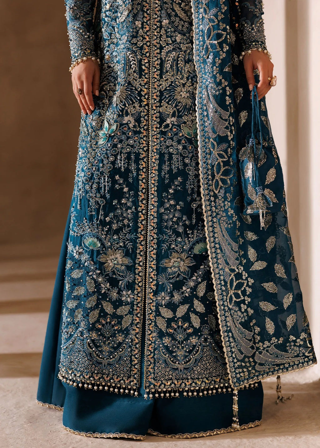 A close-up of a person wearing an ornate teal blue embroidered traditional outfit with intricate metallic detailing, holding a matching embroidered dupatta in a softly lit indoor setting.