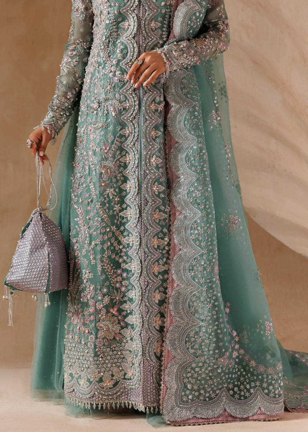 A model wears a long-sleeved mint green traditional gown with intricate silver and pink floral embroidery, holding a matching mint green clutch bag against a neutral beige background.