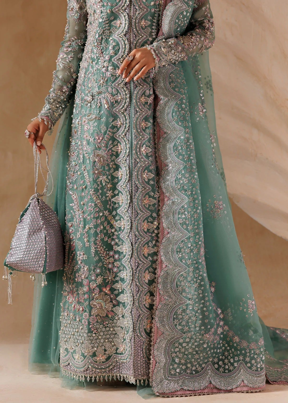 A model wears a long-sleeved mint green traditional gown with intricate silver and pink floral embroidery, holding a matching mint green clutch bag against a neutral beige background.