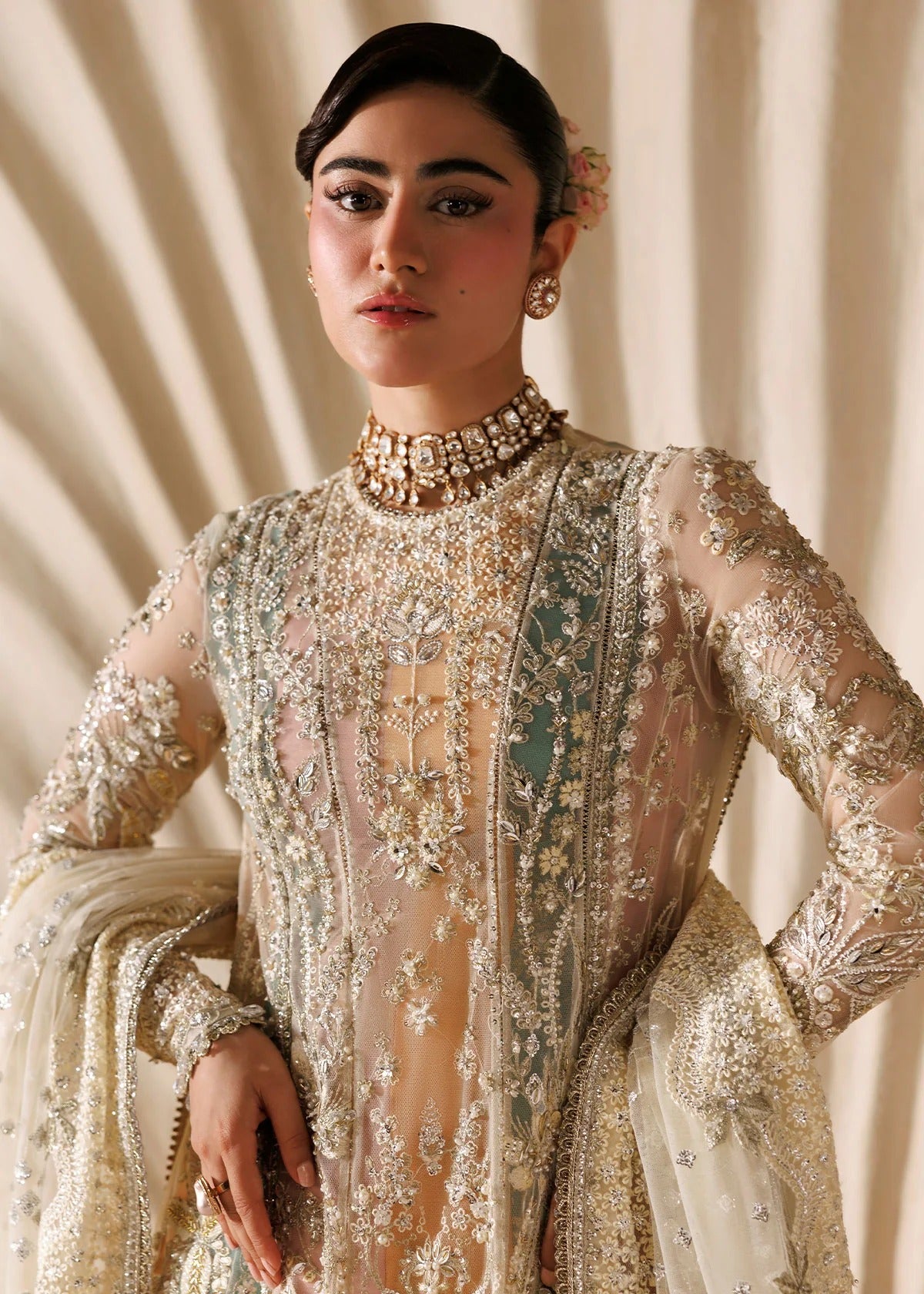 A woman modeling an ornate, heavily embellished traditional outfit with long sheer sleeves, intricate gold embroidery, and a matching draped scarf against a draped cream fabric backdrop.