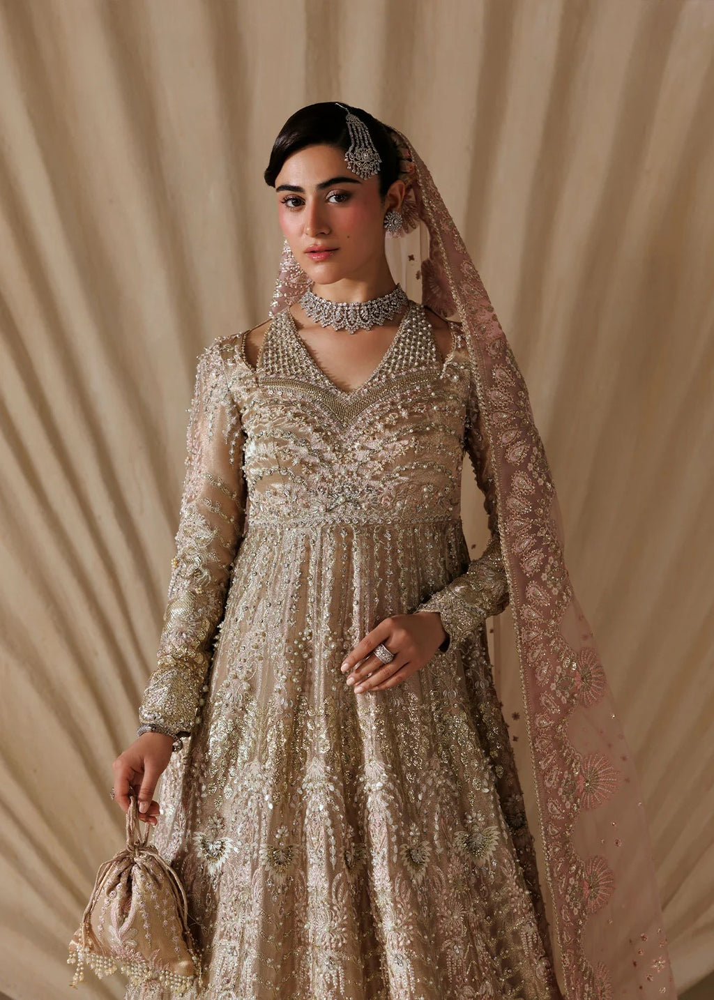 A woman models an ornate beige and gold heavily embroidered traditional dress with a matching sheer dupatta, jewelry, and a small matching handbag against a draped beige fabric backdrop.