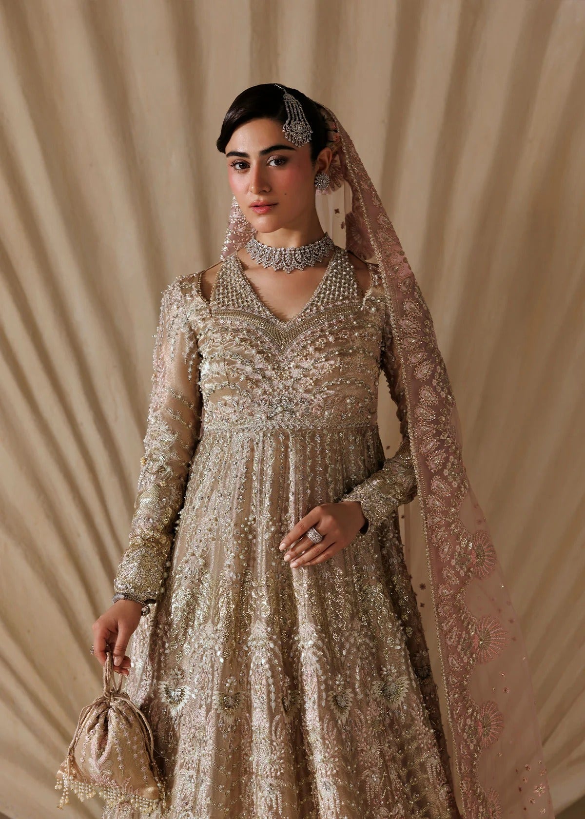 A woman models an ornate beige and gold heavily embroidered traditional dress with a matching sheer dupatta, jewelry, and a small matching handbag against a draped beige fabric backdrop.
