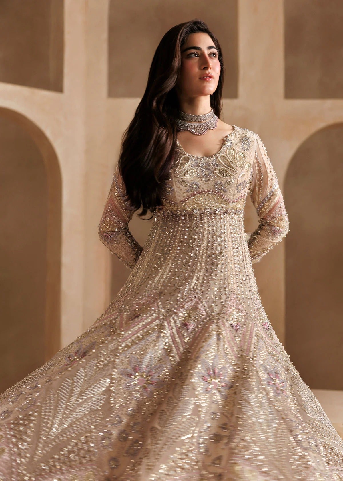 A woman modeling a heavily embellished beige and gold traditional gown with intricate embroidery, a high-neck choker, and long sheer sleeves, set against a warm beige architectural background.