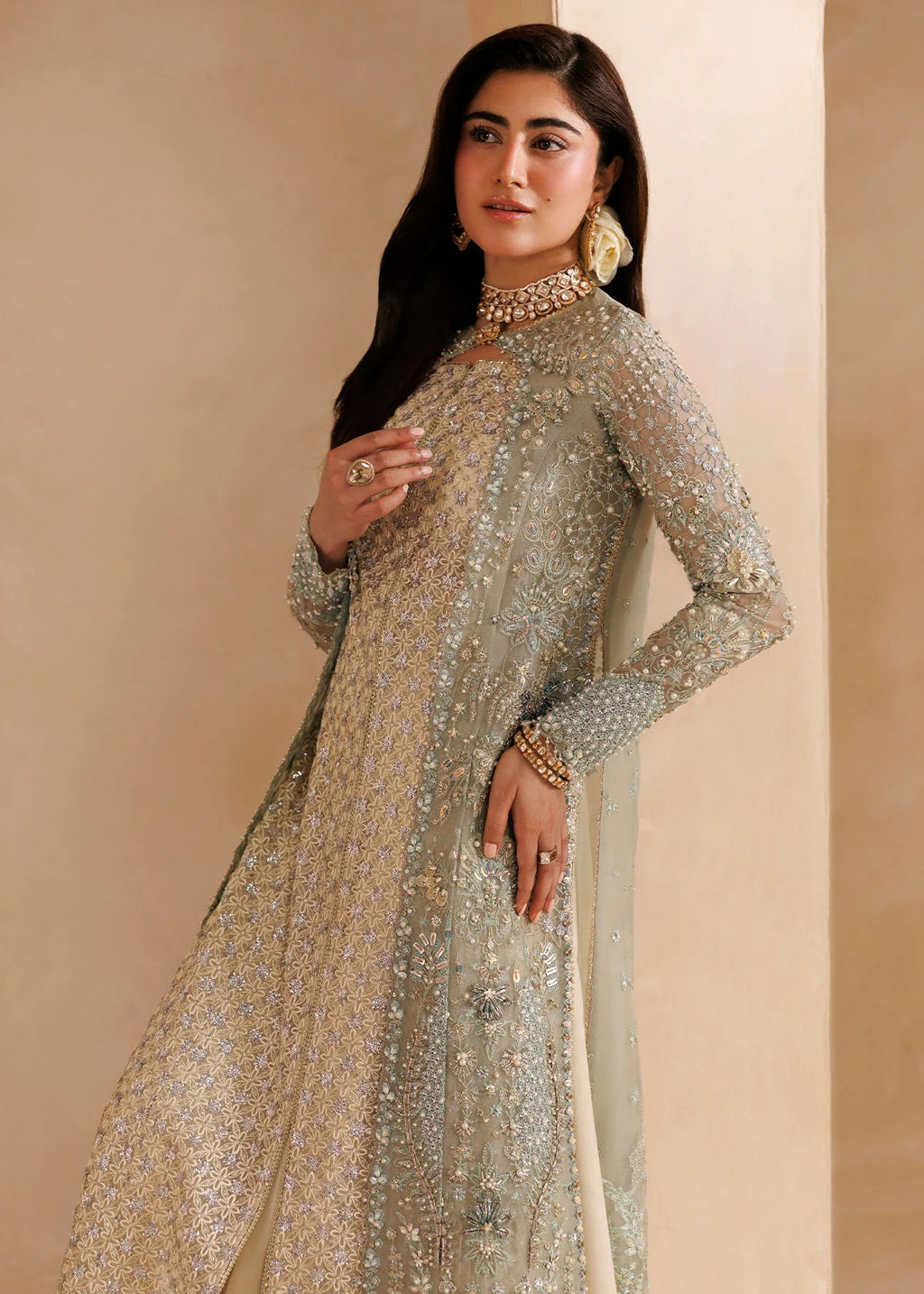 A woman modeling a long-sleeved, heavily embellished silver and gold traditional outfit with a matching sheer dupatta, set against a warm beige background.