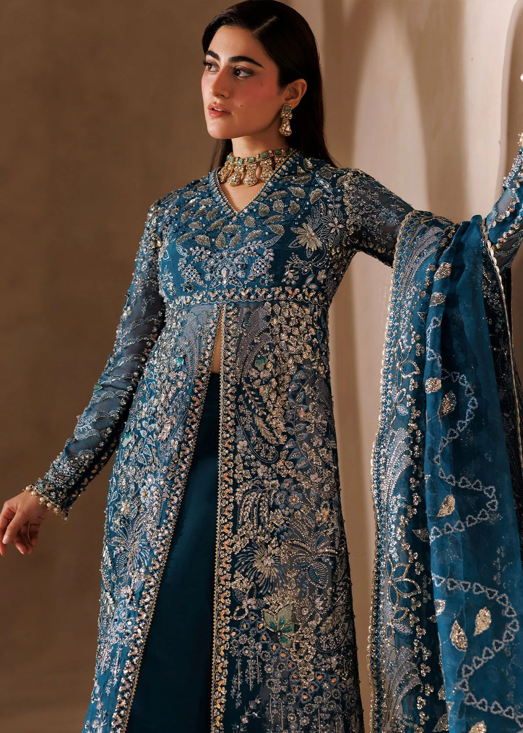 A woman models a traditional blue embroidered outfit with a long sheer jacket, matching skirt, and dupatta, decorated with intricate gold and silver mirror work and floral patterns.