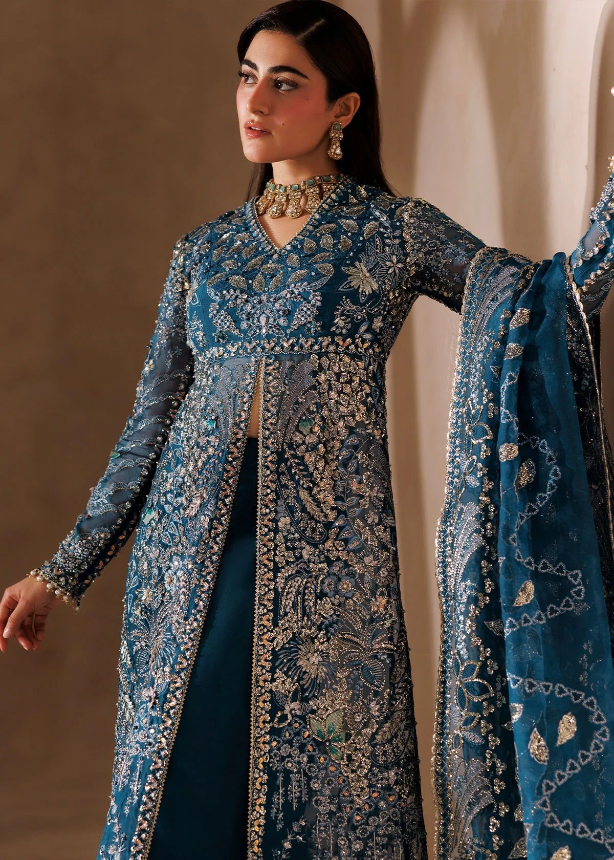 A woman models a traditional blue embroidered outfit with a long sheer jacket, matching skirt, and dupatta, decorated with intricate gold and silver mirror work and floral patterns.