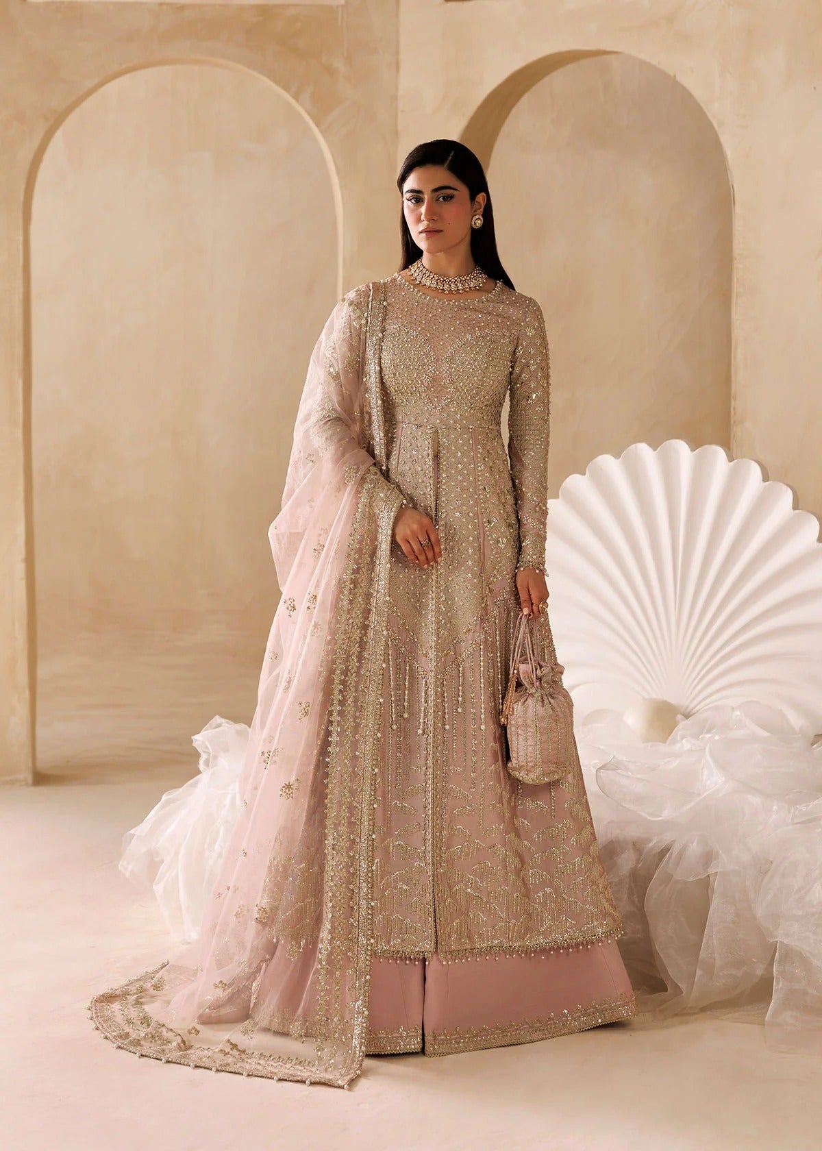 A woman models a traditional Indian anarkali suit in a warm beige studio setting with arches. The outfit is a dusty pink anarkali heavily embellished with gold embroidery and sequins, paired with a matching sheer dupatta and a white tulle skirt.