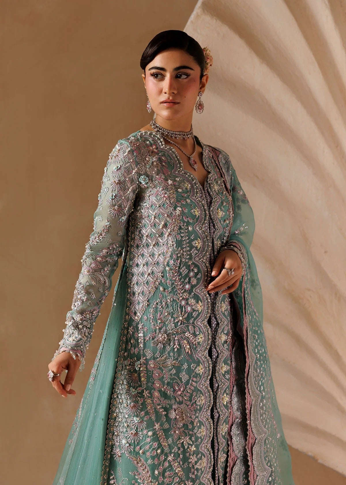 A woman modeling a traditional long-sleeve embroidered gown in mint green with intricate silver and pink floral embroidery, set against a warm beige draped backdrop.
