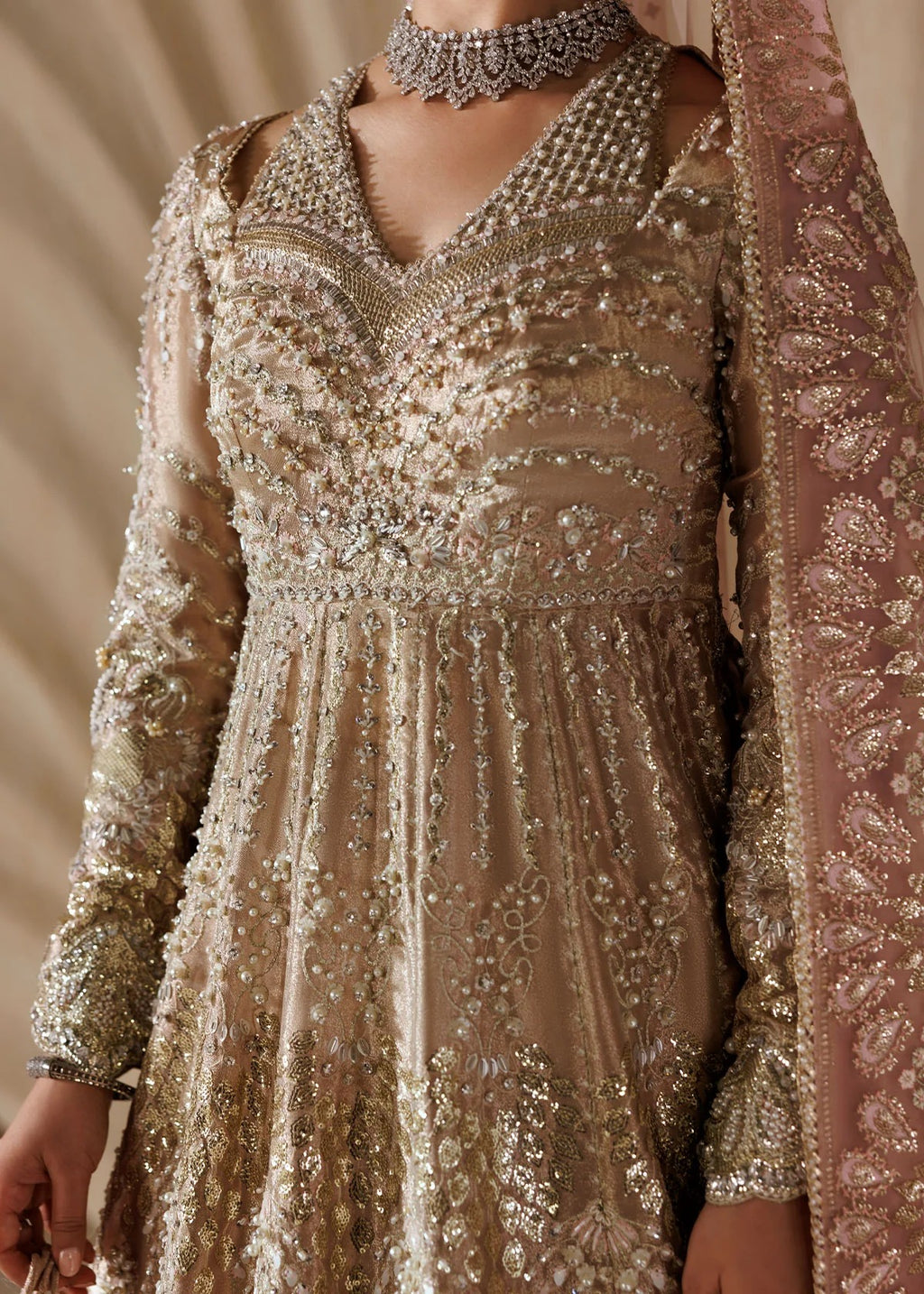 A close-up of a woman wearing an ornate beige and gold embellished traditional dress with intricate beadwork, a matching sheer dupatta, and a silver choker necklace.