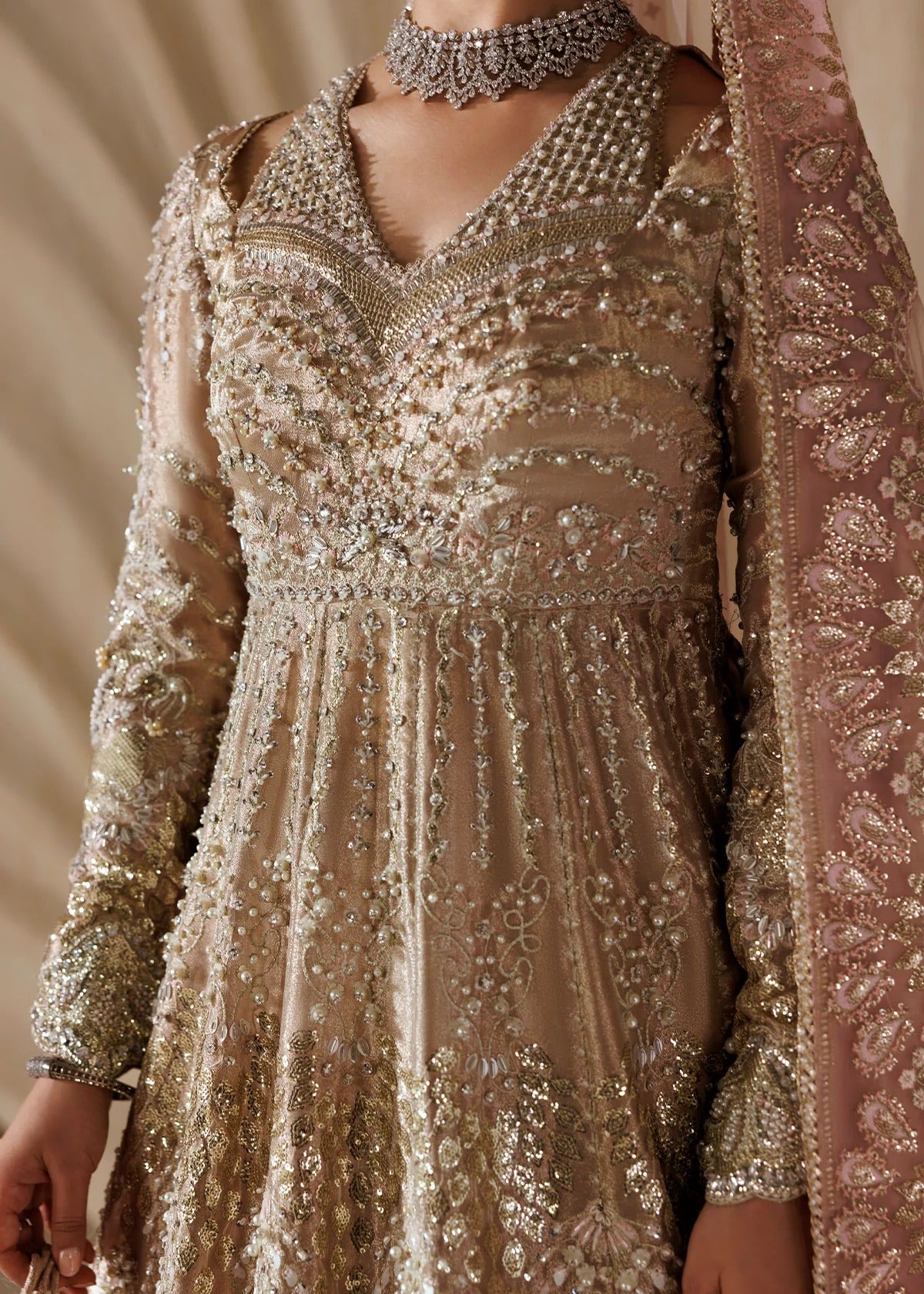 A close-up of a woman wearing an ornate beige and gold embellished traditional dress with intricate beadwork, a matching sheer dupatta, and a silver choker necklace.