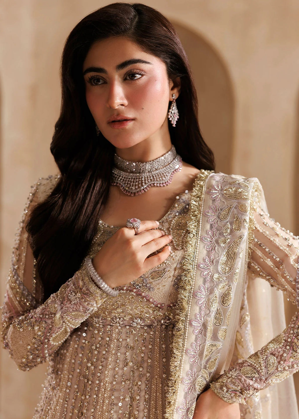 A woman modeling an ornate beige and gold embellished traditional outfit with a matching sheer dupatta, styled with a silver choker necklace and earrings against a warm beige background.