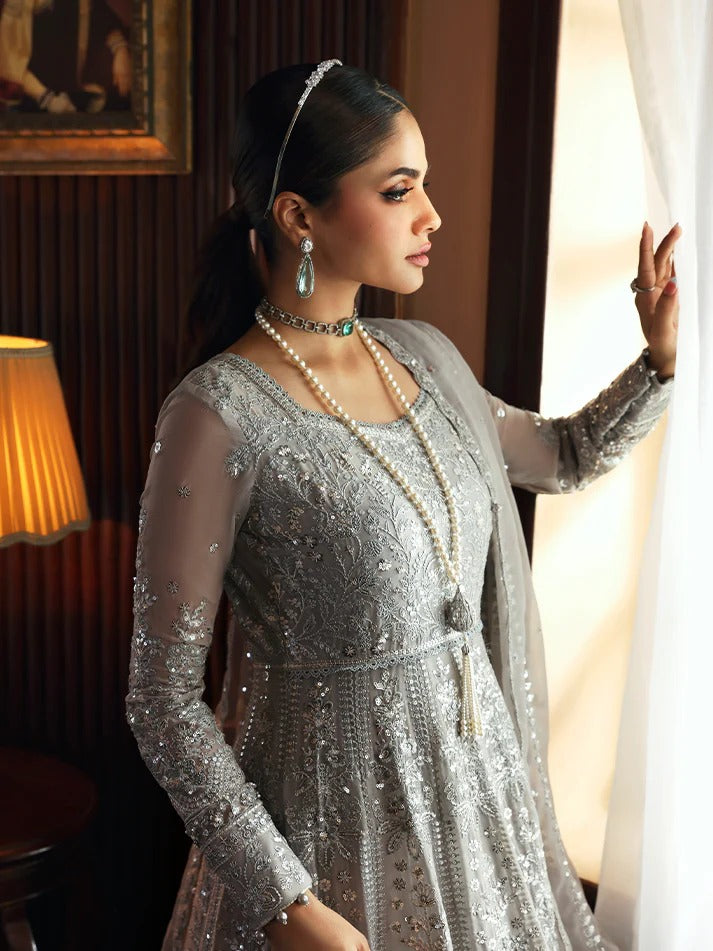 Woman in a silver embroidered outfit with jewelry, standing by a window.