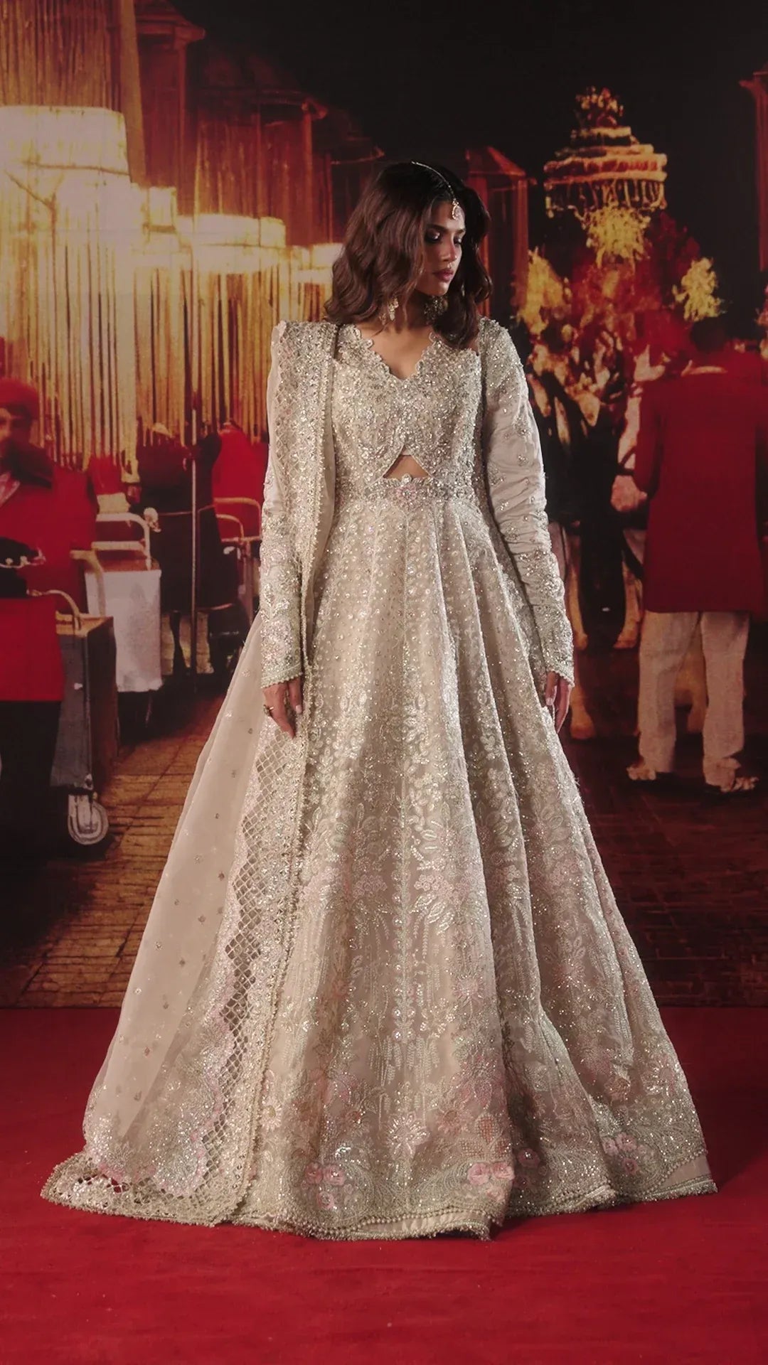 A woman wearing a heavily embellished silver and beige long-sleeve gown with a cutout at the waist, standing on a red carpet in front of a warm-toned event backdrop.