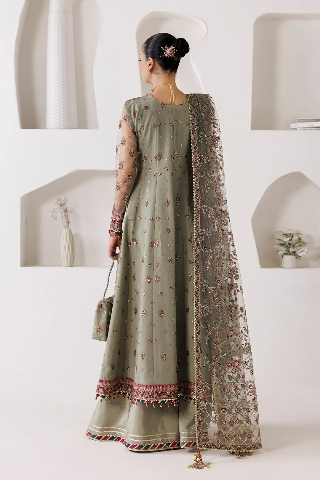 A woman wearing a sage green traditional Indian outfit with floral embroidery and a matching sheer dupatta, photographed from the back in a minimalist white interior setting.