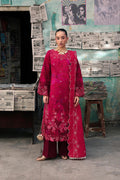 A woman stands in a rustic shop-like setting wearing a bright magenta-pink traditional outfit with floral embroidery, matching pants, and a draped scarf. Newspapers and posters cover the wall behind her.
