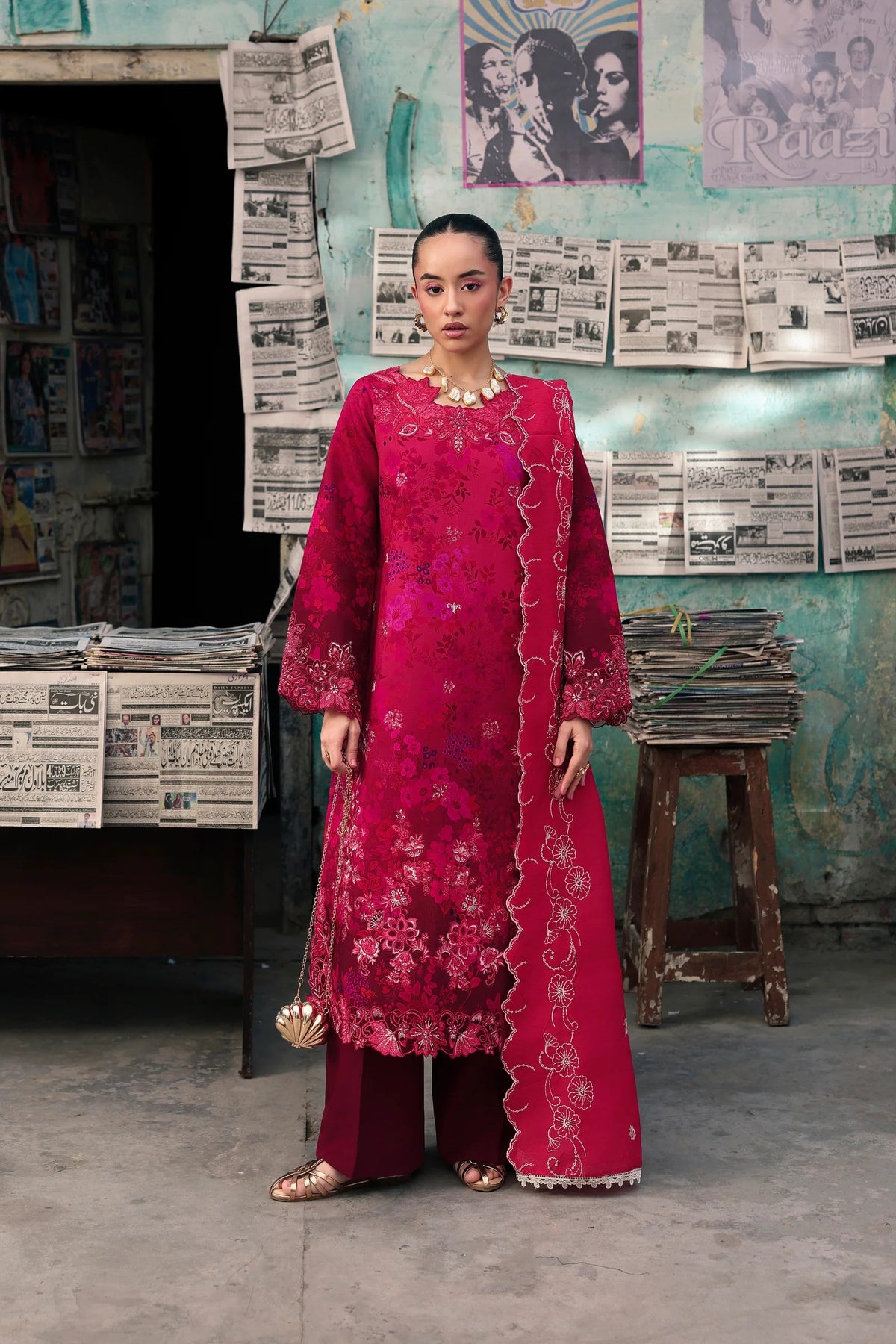 A woman stands in a rustic shop-like setting wearing a bright magenta-pink traditional outfit with floral embroidery, matching pants, and a draped scarf. Newspapers and posters cover the wall behind her.