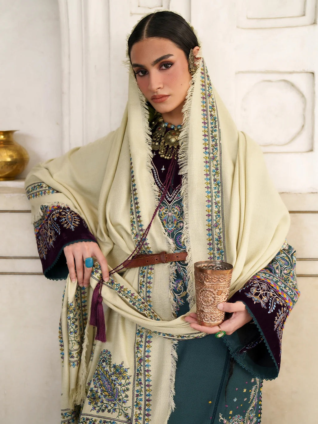 A woman models an intricate traditional outfit featuring a cream-colored shawl with colorful embroidery, a deep purple embroidered tunic, and a teal skirt, holding a carved wooden cup against an elegant white paneled background.