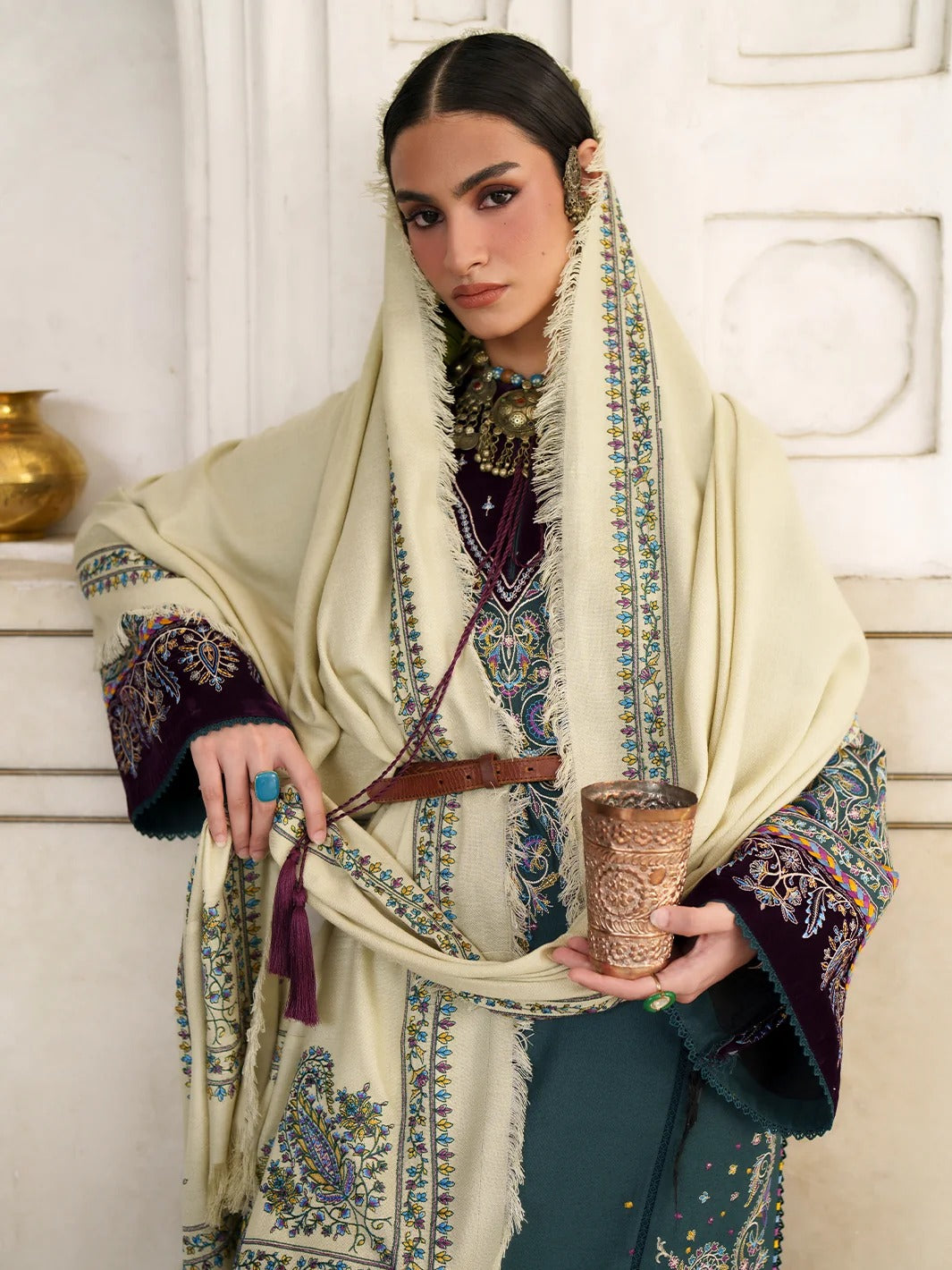 A woman models an intricate traditional outfit featuring a cream-colored shawl with colorful embroidery, a deep purple embroidered tunic, and a teal skirt, holding a carved wooden cup against an elegant white paneled background.