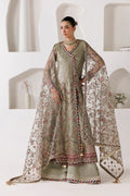 A woman models a traditional South Asian salwar kameez suit in sage green and beige with intricate floral embroidery and a matching sheer dupatta, set against a modern white wall with built-in arches.