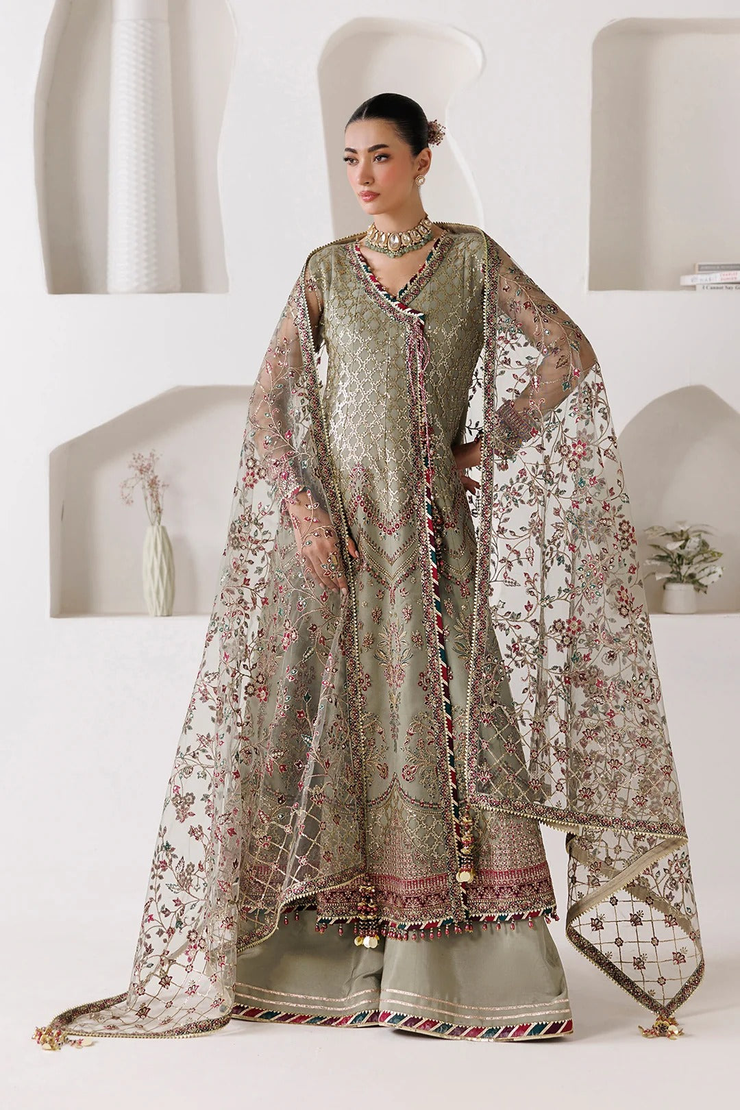 A woman models a traditional South Asian salwar kameez suit in sage green and beige with intricate floral embroidery and a matching sheer dupatta, set against a modern white wall with built-in arches.