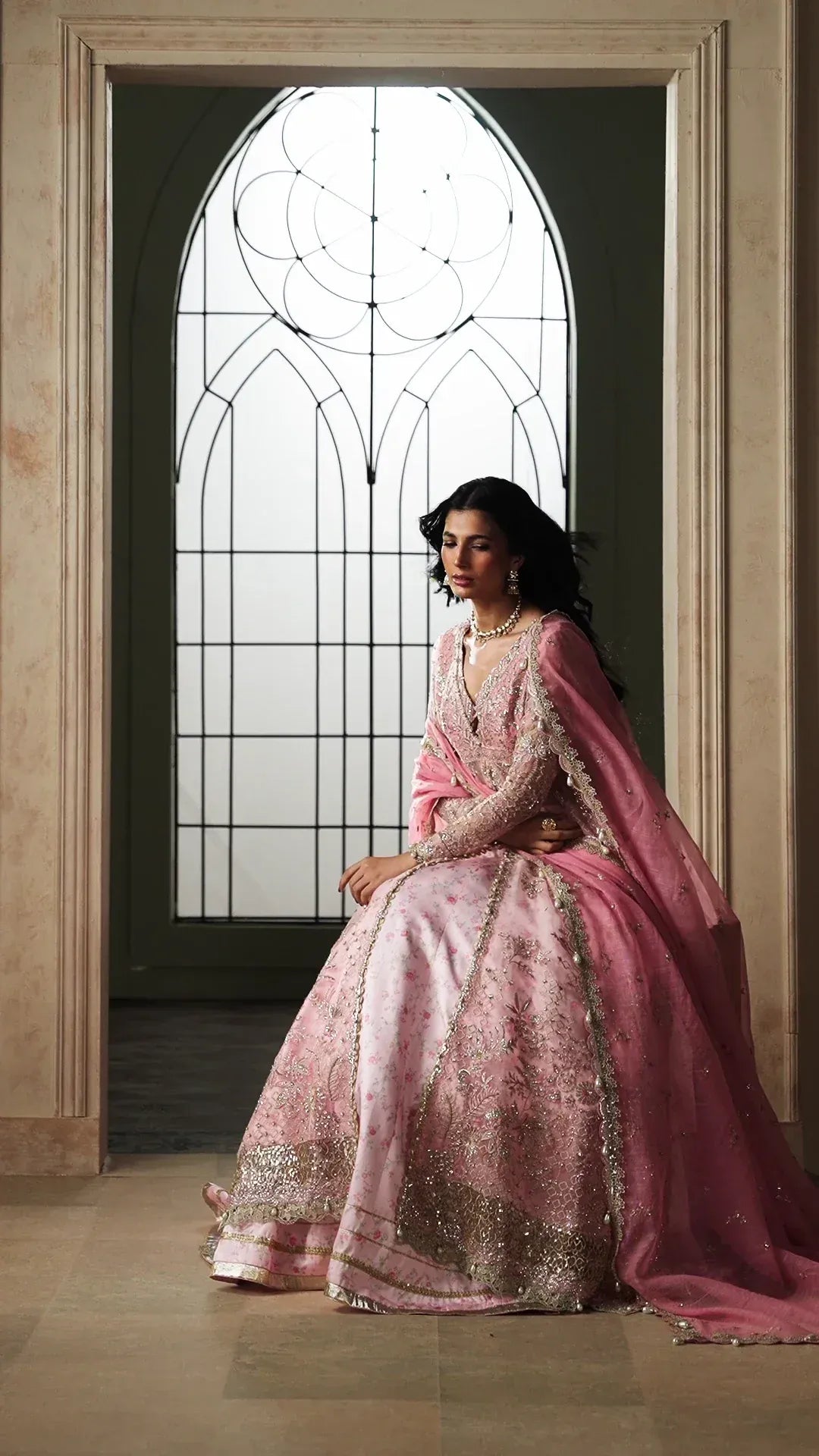 A woman wearing a pink embroidered traditional gown with a matching dupatta sits in a grand arched doorway with a stained-glass window behind her.