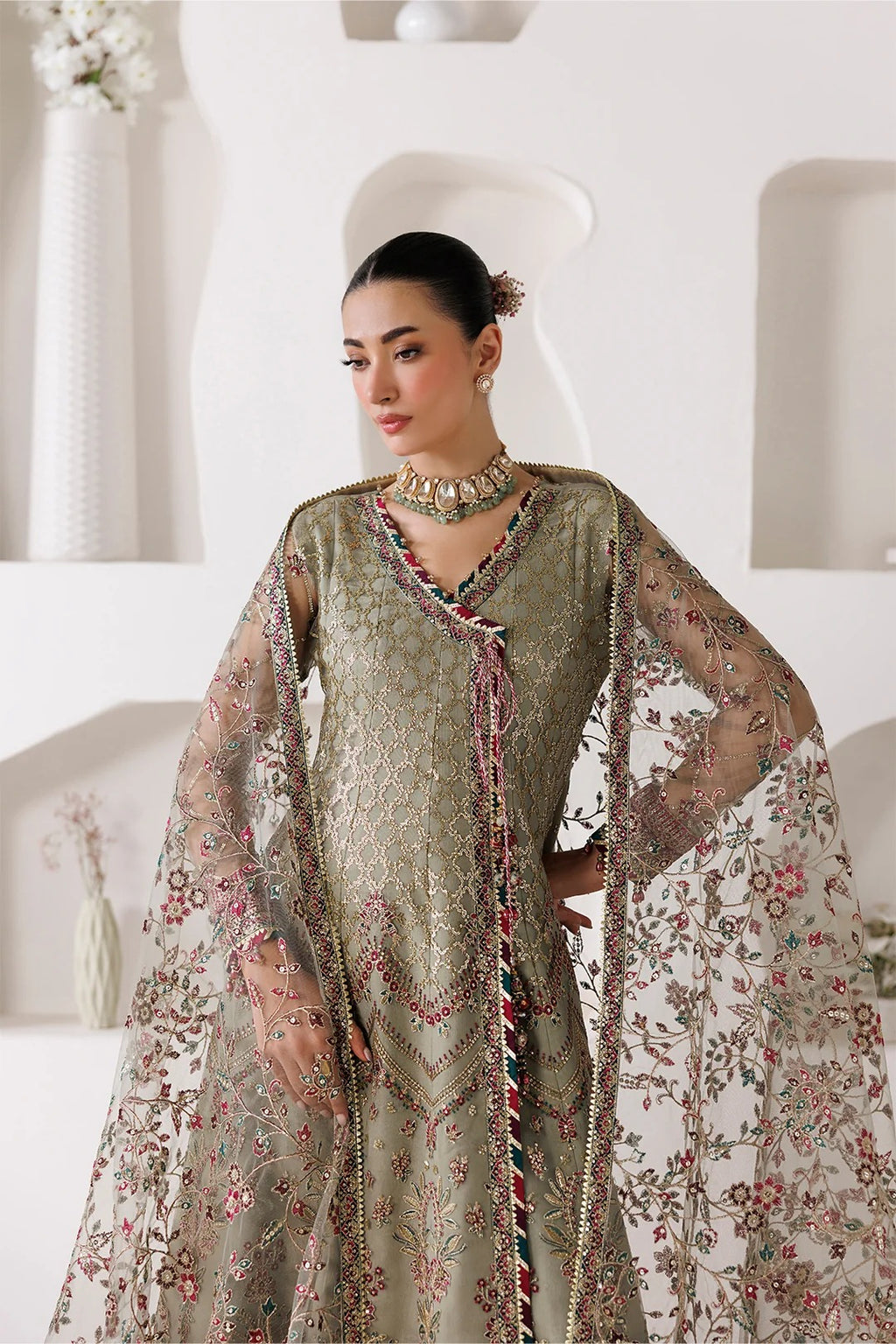 A woman models a traditional South Asian outfit featuring a heavily embroidered olive green kurta with a matching sheer floral dupatta, set against a clean, modern white architectural background.