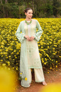 A woman stands in a field of yellow flowers wearing a traditional three-piece salwar kameez outfit in light sage green and cream with intricate embroidery, a matching sheer jacket, and wide-leg trousers.