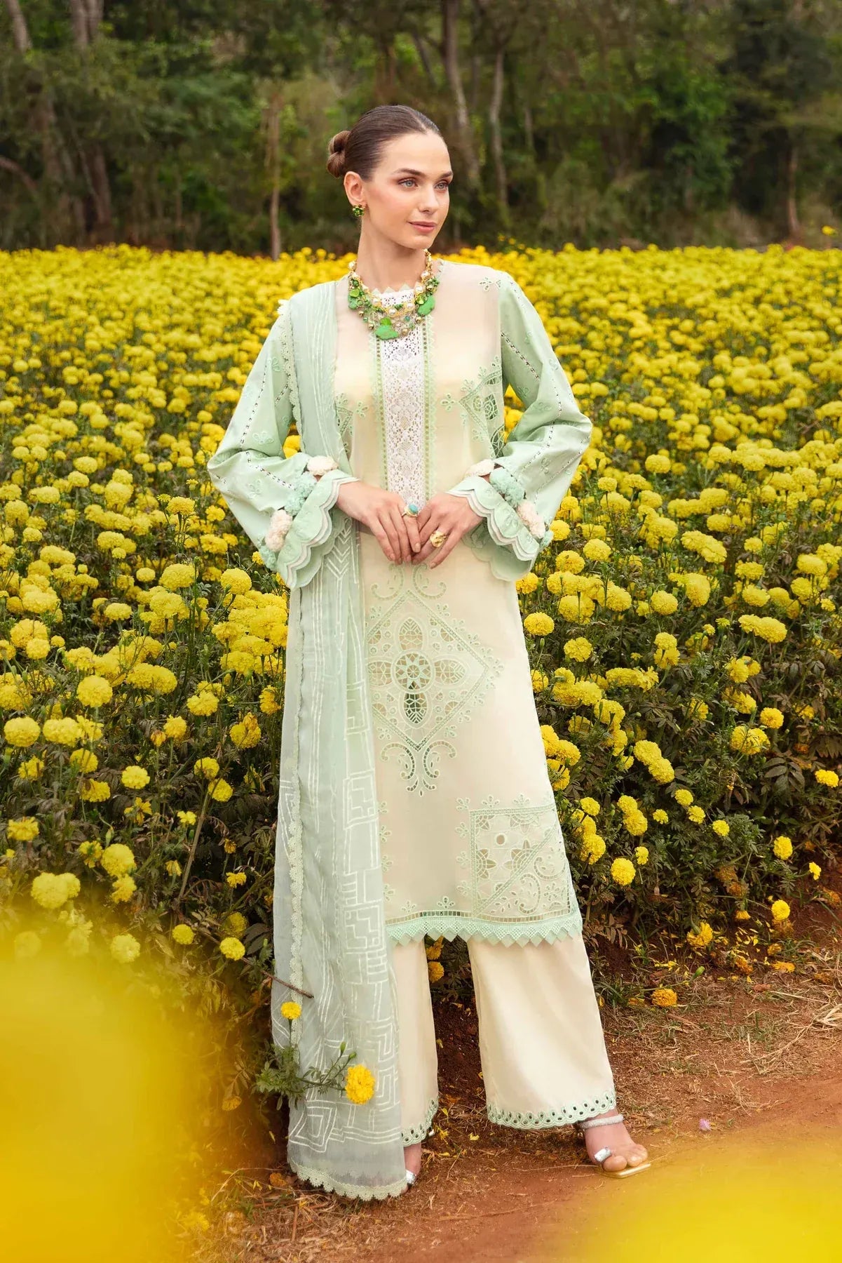 A woman stands in a field of yellow flowers wearing a traditional three-piece salwar kameez outfit in light sage green and cream with intricate embroidery, a matching sheer jacket, and wide-leg trousers.