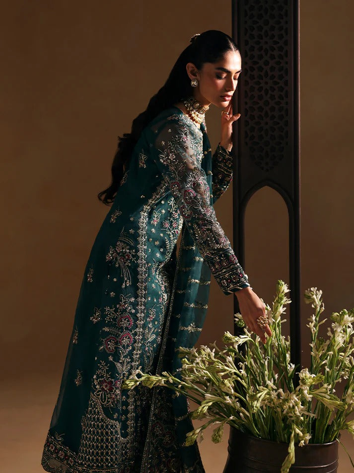 Woman in a teal embroidered outfit holding a plant against a warm-toned wall.
