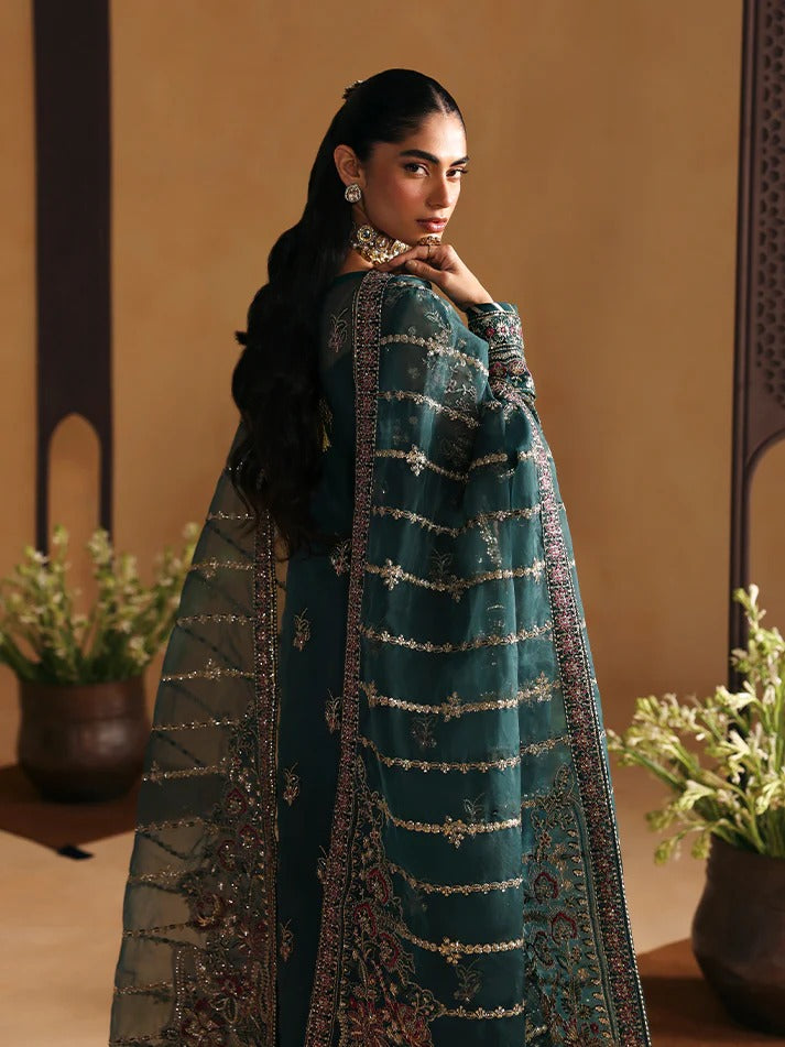 Woman in a traditional outfit with intricate patterns, standing against a beige wall with plants.