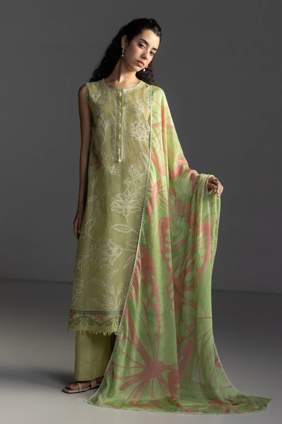 A woman models a traditional three-piece salwar kameez suit in sage green with white floral embroidery, paired with a matching sheer dupatta featuring pink and green abstract patterns, set against a dark gray studio background.